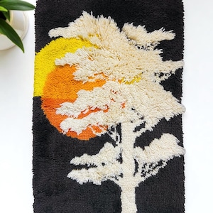 May include: A textured wall hanging featuring a stylized tree and sunset design. The artwork uses a black background with a yellow, orange, and white color palette. The tree is white, and the sun is a gradient of yellow and orange.