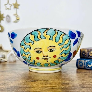 May include: A white ceramic bowl featuring a hand-painted yellow sun face with a teal wavy border. The bowl is decorated with blue and orange accents around the rim and sides, showcasing a bright, colorful design on a rounded, deep vessel.
