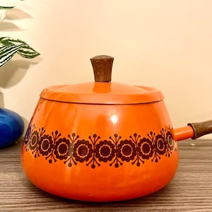 May include: Orange enamel fondue pot with a brown wooden handle and a floral design around the rim. The pot has a lid with a brown wooden knob.