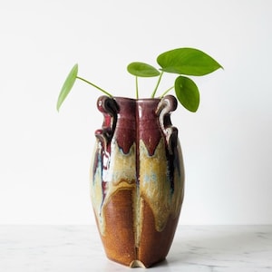 May include: A handcrafted ceramic vase with a unique shape and a blend of colors. The vase features a dark burgundy top, transitioning to a warm brown base with dripped glaze effects. Green plant stems and leaves emerge from the vase.
