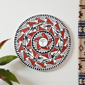 May include: Round decorative plate with a vibrant geometric design in red, black, and white. The plate features a central design with the text "Costa Rica." The plate is mounted on a white wall.