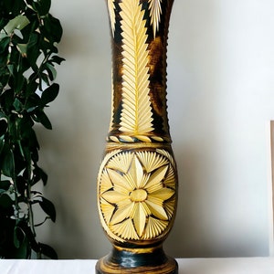 Könnte beinhalten: Eine hohe, handgeschnitzte Holzvase mit aufwendigen Blumen- und Blattmotiven. Die Vase hat eine dunkelbraune und natürliche Holzfarbe, mit einem breiten Sockel und einem ausgestellten oberen Teil. Die detaillierten Schnitzereien verleihen einen rustikalen, künstlerischen Touch.