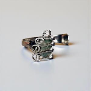 May include: A silver-toned ring with a unique design. The ring features a series of swirling, scroll-like elements and a rectangular section with a green hue. The band is simple, and the overall aesthetic is artistic and modern.