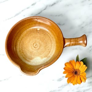 May include: A brown ceramic skillet with a single handle. The skillet has a light brown glaze and a slightly textured surface.