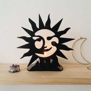 May include: A black metal candle holder shaped like a sun with a stylized face, featuring sharp, pointed rays extending outward. A small, lit tea light candle is positioned in the center, casting a warm glow through the cutout design.