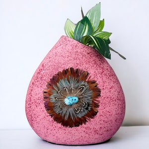May include: A pink ceramic vase with a speckled finish and a decorative feather and turquoise stone design. The vase is shaped like a teardrop and has a small opening at the top. A small green plant is in the vase.