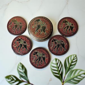 May include: A set of six decorative coasters and a round box. The coasters and box are black with red and green elephant designs. The coasters are round, and the box has a lid. The coasters and box are on a white marble surface.