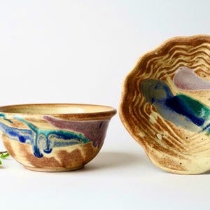 May include: Two ceramic bowls with a cream-colored base and wavy brown textures. One bowl is upright, showing blue and purple glaze drips on the exterior. The other bowl is tilted, revealing a blue and purple abstract design inside.