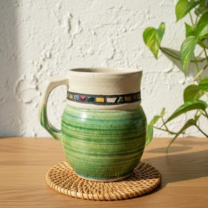 May include: A handmade ceramic mug with a cream-colored rim and handle, and a green body with horizontal stripes. The mug features a decorative band with colorful squares. It sits on a round woven coaster.
