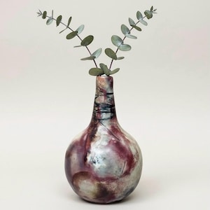 May include: A handcrafted ceramic vase with a bulbous body and a narrow neck. The vase features a blend of colors, including burgundy, silver, and black, with a glossy finish. Two sprigs of eucalyptus are arranged in the vase, adding a touch of greenery.