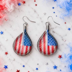 Könnte beinhalten: Ein Paar tropfenförmige Ohrringe mit einem gewickelten Metalldrahtrahmen, der mit rotem, weißem und blauem Garn umwickelt ist, um ein Muster zu erzeugen, das an die amerikanische Flagge erinnert. Die Ohrringe haben silberfarbene Metallhaken.