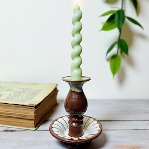 May include: A lit, light green, spiral-shaped candle sits in a brown and white ceramic candlestick holder. The candlestick has a round base with a decorative white and brown pattern. The candle is lit and has a small flame.