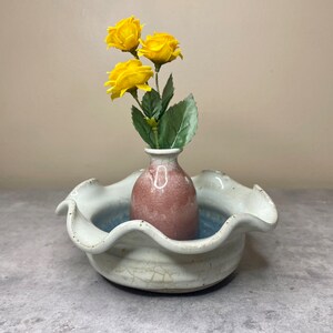 May include: A white ceramic bowl with a ruffled edge, featuring a small pink vase with yellow flowers inside. The bowl has a crackled glaze and a blue glaze on the inside.