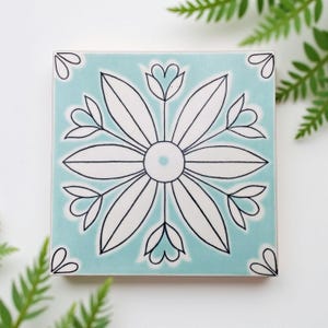 May include: Square ceramic tile with a light blue background and a large white flower design outlined in black. The flower has multiple petals and a central circle. Small heart-shaped designs are in each corner.