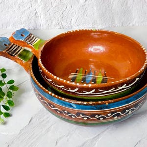 May include: Two terracotta bowls with blue and green painted designs. The smaller bowl has a handle and is nested inside the larger bowl.