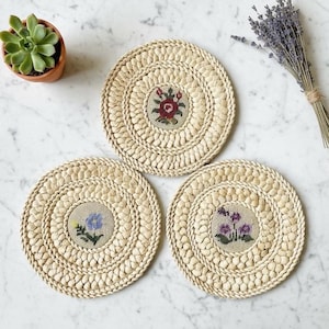 May include: Three round woven coasters with embroidered floral designs in red, blue, and purple. The coasters are made of natural fibers and have a textured, braided edge. A small potted succulent and a bundle of lavender are also in the image.