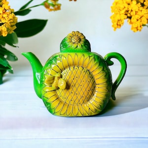 May include: A ceramic teapot with a sunflower design. The teapot is primarily green and yellow, with a large sunflower painted on the front and a smaller sunflower on the lid. A bee is sculpted on the side. The teapot is on a white surface.