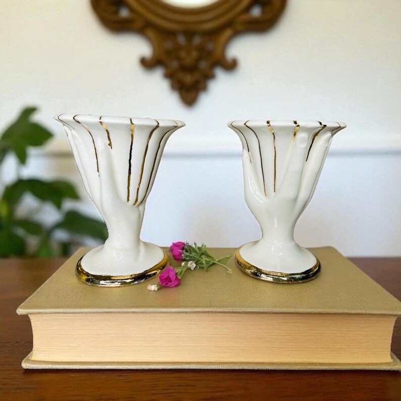 K&ouml;nnte beinhalten: Zwei wei&szlig;e Keramikvasen mit handf&ouml;rmigem Design und goldenen Details. Die Vasen haben goldene Streifen und stehen auf einem Buch. Das Bild zeigt auch einen dekorativen Spiegel und einige rosa Blumen.