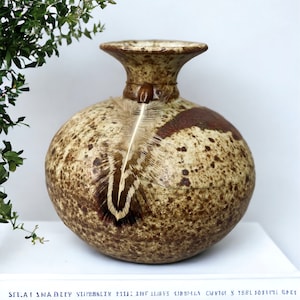 May include: A speckled brown ceramic vase with a wide mouth and a feather inside. The vase has a mottled brown and white glaze.