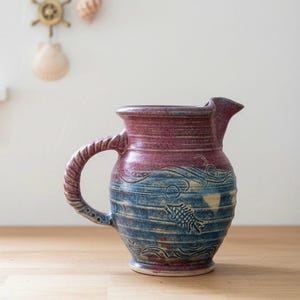 Peut inclure: Pichet en céramique fait à la main avec une anse torsadée. Le haut est rouge foncé, passant au bleu avec un motif de poisson et de vagues. Le pichet mesure environ 20 cm de haut et est une pièce unique de décoration de cuisine.