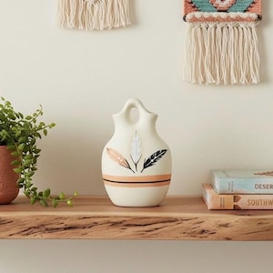 May include: A cream-colored ceramic vase with a double-spout handle design. The front features three painted feathers in peach, white, and black, positioned above two horizontal lines in peach and black that wrap around the lower body.