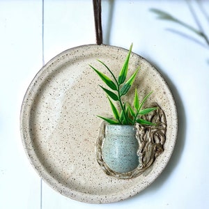 May include: A white ceramic wall planter with a speckled finish. The planter has a small, round opening for a plant and a rope-like design around the opening. A small green plant is growing out of the planter.