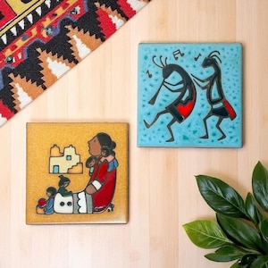 May include: Two square ceramic tiles with Native American designs. One tile shows a woman with children in a red dress, the other depicts two figures playing music on a turquoise background. The tiles are on a light wood surface.