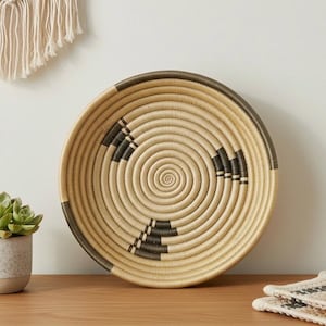 May include: A round, woven basket with a spiral design. The basket features a natural beige color with dark brown accents. The rim and sections of the design are dark brown. The basket is on a wooden surface.