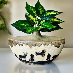 Könnte beinhalten: Eine Keramikvase mit einem gewellten Rand und einer cremefarbenen Glasur. Die Vase zeigt schwarze Silhouetten eines Hirsches, von Bäumen und einem Vogel. Die Vase ist mit grünen, blättrigen Pflanzen gefüllt.