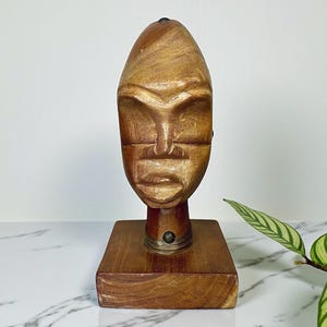 May include: A carved wooden sculpture of a stylized face. The face is light brown with carved features, mounted on a square wooden base. A metal collar encircles the neck. The sculpture is displayed on a white and gray marble surface.