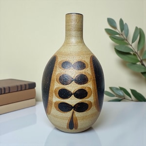 May include: A ceramic vase with a beige base and a brown and black pattern. The vase has a narrow neck and a rounded body. The design features a series of stylized, symmetrical shapes. The vase is a decorative item.
