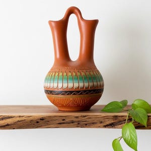 May include: A terracotta-colored pottery vase with a unique double-spout design. The vase features a decorative band of turquoise, green, and black geometric patterns around the middle. The base has carved triangular and linear designs. It sits on a wooden shelf.