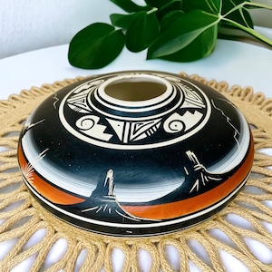 May include: Black and white pottery bowl with a geometric design and a red and white band around the rim. The bowl is sitting on a woven mat.