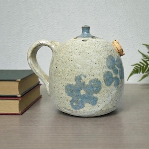 May include: A speckled ceramic teapot with a handle, spout, and lid. The teapot is decorated with blue floral designs. A cork stopper is inserted into the side. Two books are in the background.