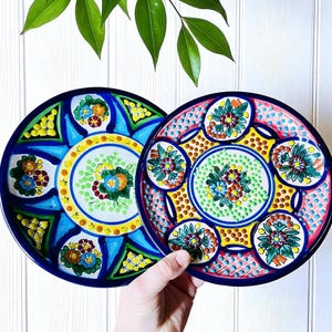 May include: Two hand-painted ceramic plates with vibrant floral designs. One plate features a blue and yellow color scheme, while the other showcases a pink and yellow palette. Both plates have a dark blue border and are decorated with colorful flowers.