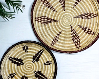 May include: Two round woven baskets. The baskets are made of natural-colored fibers with dark brown accents and rims. The larger basket has a spiral design with radiating lines, while the smaller one has a similar pattern. The baskets are on a white surface.