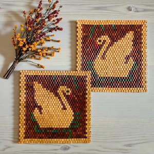 May include: Two square, woven trivets with a swan design. The swans are light tan, set against a dark brown background, and framed with a light tan border. Green and red accents add detail. A bundle of dried flowers sits nearby.
