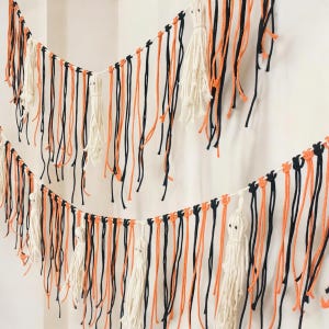 May include: Halloween garland with black, orange, and white yarn strands. The banner includes white tassel ghosts. The decorative piece is strung on a white cord, ideal for festive holiday decorations. The garland is a seasonal decoration.