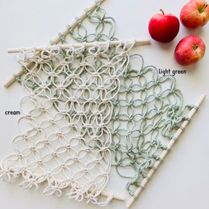 May include: A macrame wall hanging with a cream and light green color scheme. The macrame is made with a knotting technique and is attached to two wooden dowels.