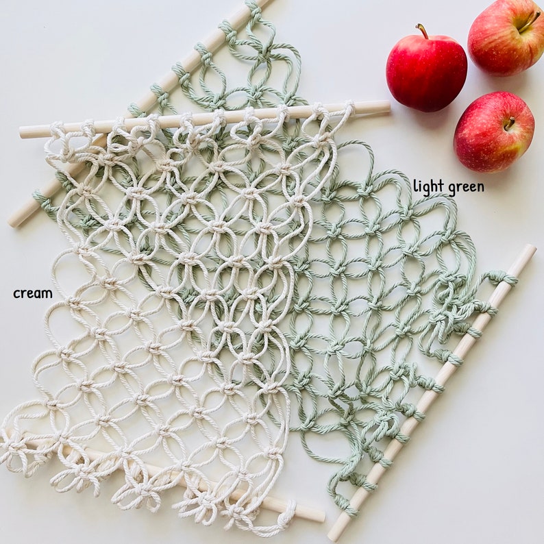 May include: A macrame wall hanging with a cream and light green color scheme. The macrame is made with a knotting technique and is attached to two wooden dowels.