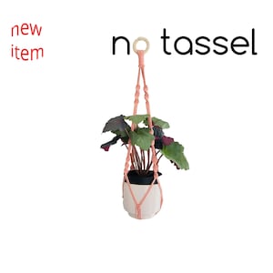 May include: A pink macrame plant hanger with a wooden ring at the top. The hanger is holding a white pot with a plant inside. The text "no tassel" is written above the hanger.