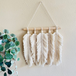 May include: A macrame wall hanging with five white feather shapes and wooden beads. The hanging is attached to a wooden dowel rod.