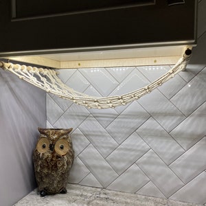 May include: A white rope hammock hangs from a wooden bar attached to a white tiled wall. A brown ceramic owl figurine sits on the floor below the hammock.