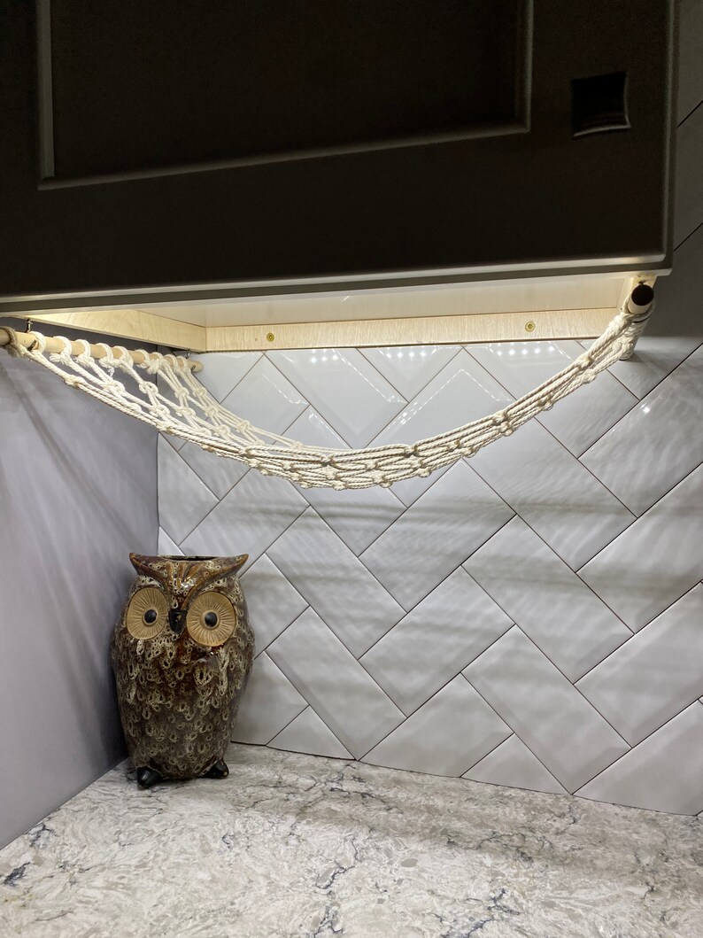 May include: A white rope hammock hangs from a wooden bar attached to a white tiled wall. A brown ceramic owl figurine sits on the floor below the hammock.