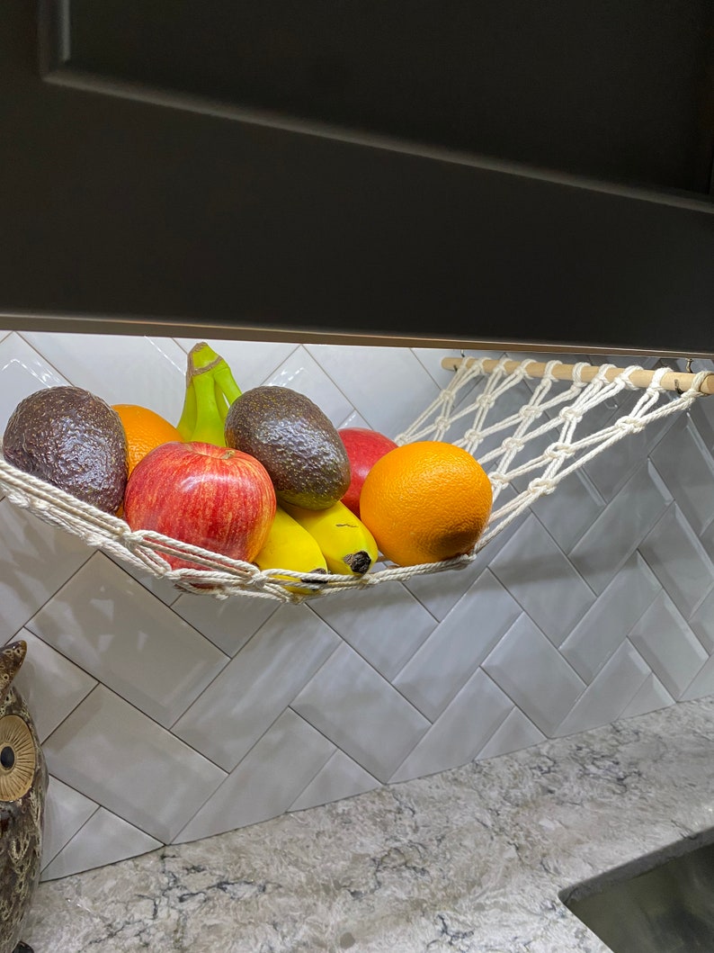 May include: A white rope hammock hanging from a cabinet, filled with fruit. The hammock holds two avocados, a red apple, two bananas, and an orange.