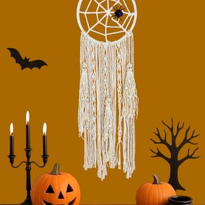 Peut inclure: Une décoration d'Halloween avec un attrape-rêves en macramé blanc, une araignée, des citrouilles, un chandelier et une silhouette de chauve-souris sur fond orange. La citrouille sculptée a un visage de lanterne.