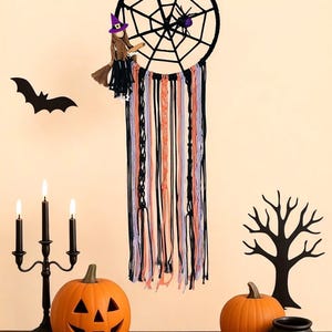 May include: A Halloween-themed display featuring a black dreamcatcher with a spider and witch, orange pumpkins, a black candelabra with lit candles, a black cauldron, and a bat and tree silhouette. The dreamcatcher has colorful hanging yarn.