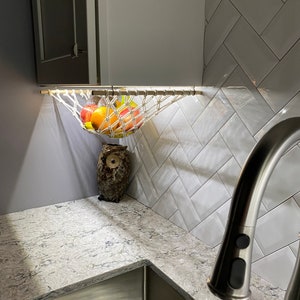 May include: A white string hammock hanging from a cabinet above a kitchen sink. The hammock is filled with fruit, including apples, oranges, and lemons. A ceramic owl figurine sits on the counter below the hammock.