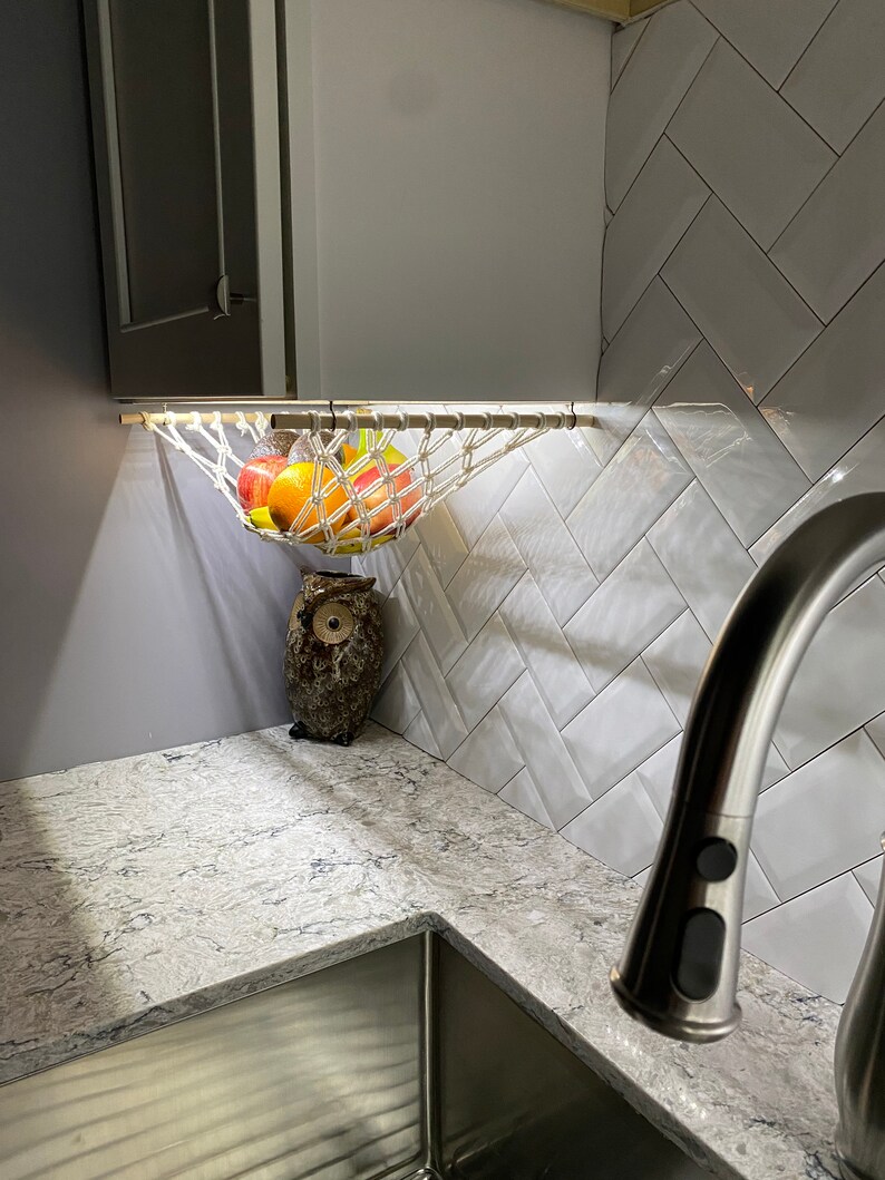 May include: A white string hammock hanging from a cabinet above a kitchen sink. The hammock is filled with fruit, including apples, oranges, and lemons. A ceramic owl figurine sits on the counter below the hammock.