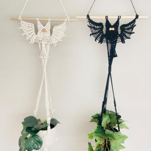 May include: Two macrame bat plant hangers, one white and one black, suspended from wooden dowels. Each bat has outstretched wings and holds a white or black pot with greenery. A decorative and functional home decor item.